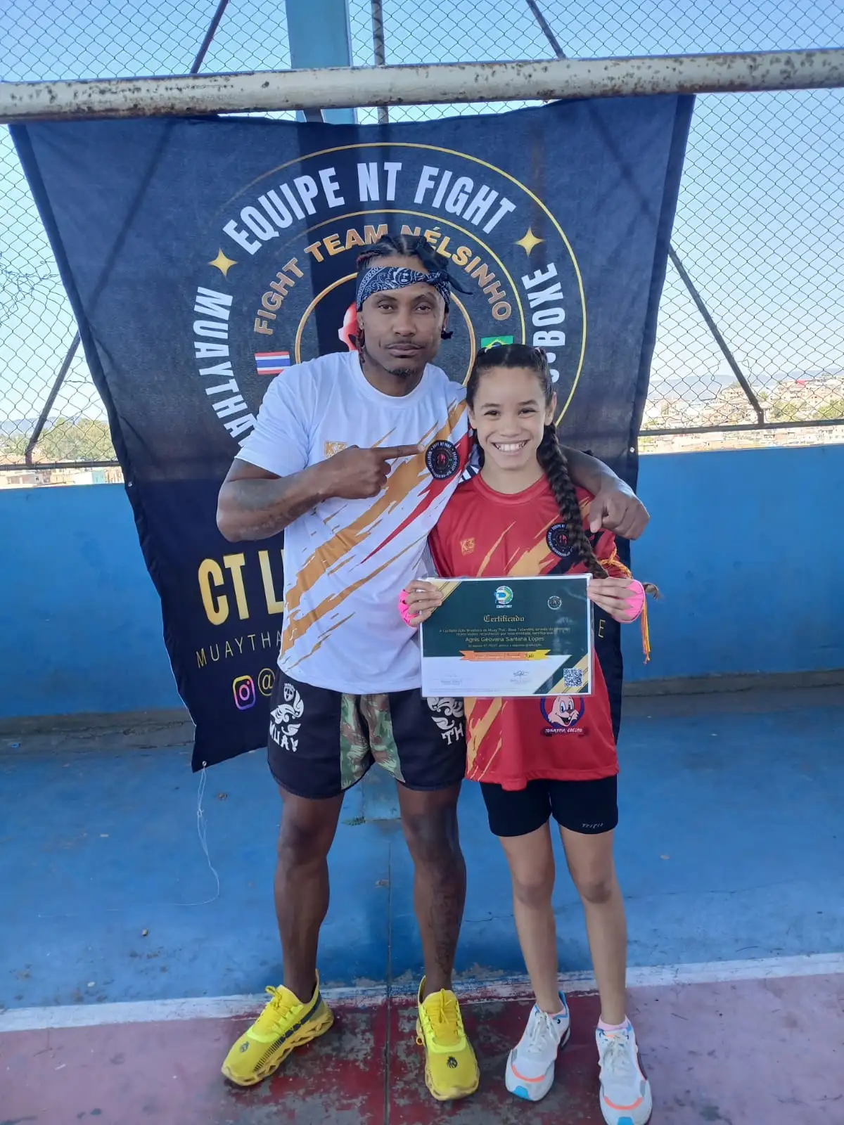 Aluna da turma kids segurando o certificado ao lado do Prof. Nélsinho, diante da bandeira da Equipe NT Fight