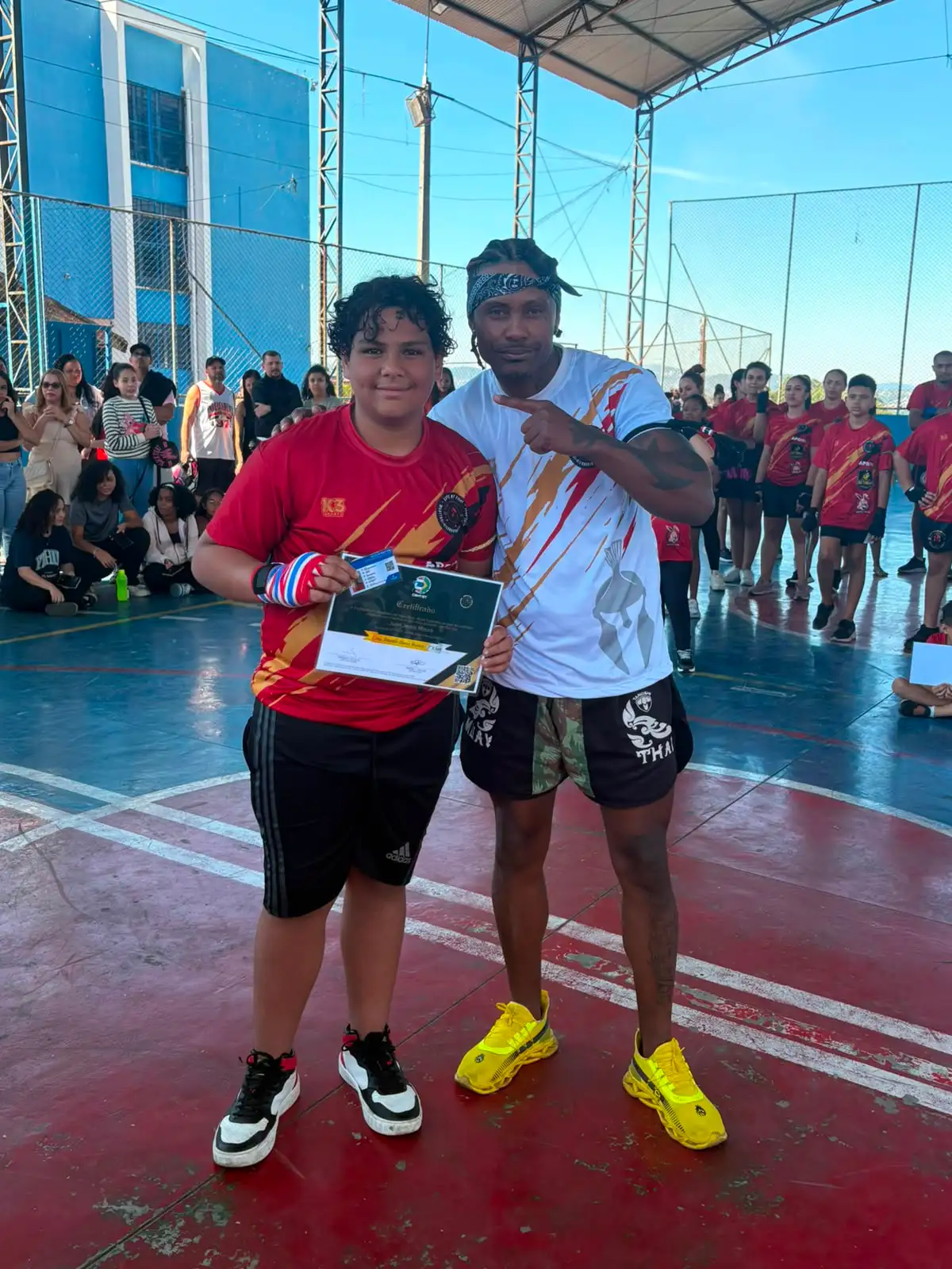 Aluno da Equipe NT Fight com o certificado de graduação ao lado do Prof. Nélsinho, na cerimônia de julho de 2026